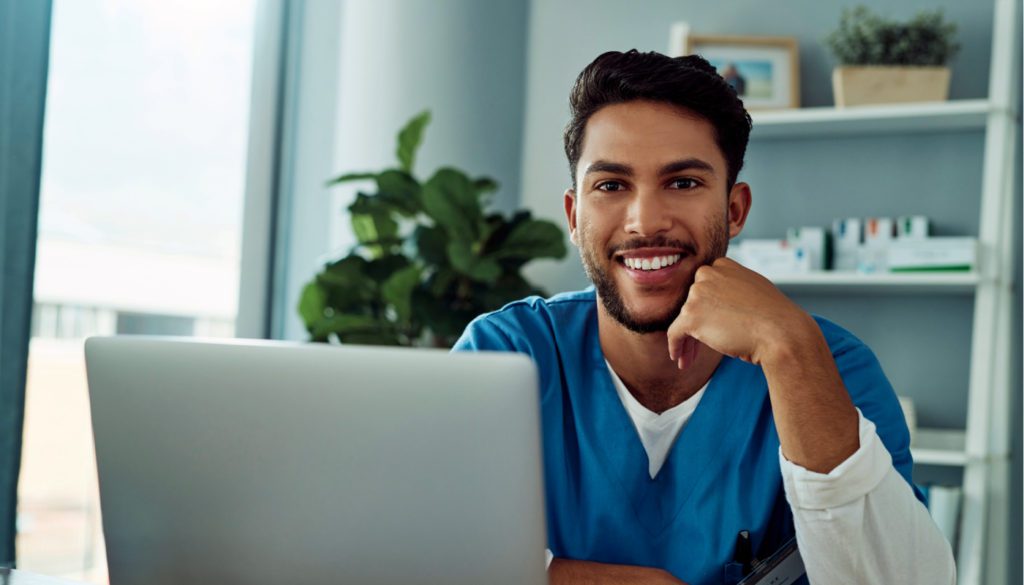 image: man in medical scrubs smiling at camera in front of his computer