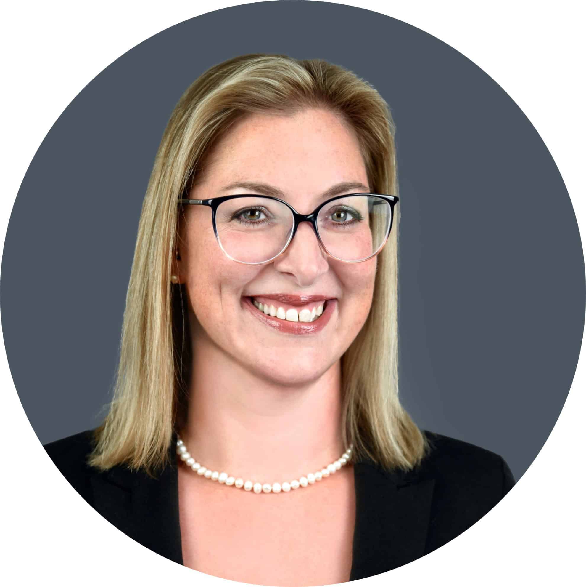 A woman with shoulder-length blonde hair, wearing glasses, a black blazer, and a pearl necklace, smiles at the camera against a gray background.