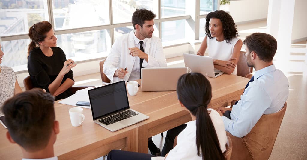 Six people in business attire seated around a table with laptops and notepads, engaged in a discussion in a bright office setting.