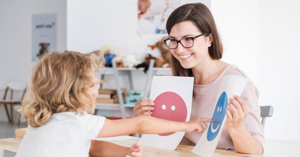 A woman shows a child two smiley face cards. The child points to the card with a blue smiley face.