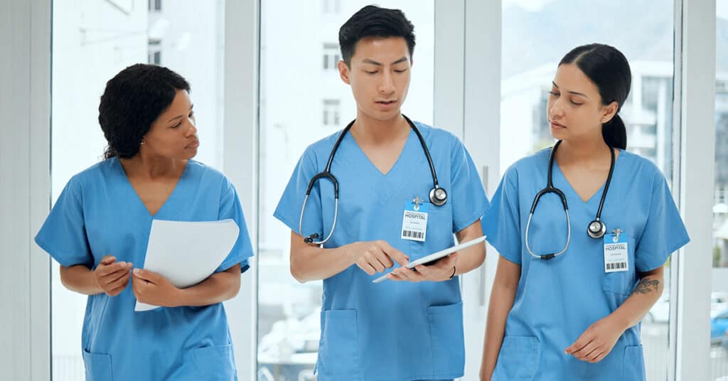 Three healthcare professionals in blue scrubs stand indoors, reviewing information on a tablet and holding documents, with stethoscopes and ID badges visible.