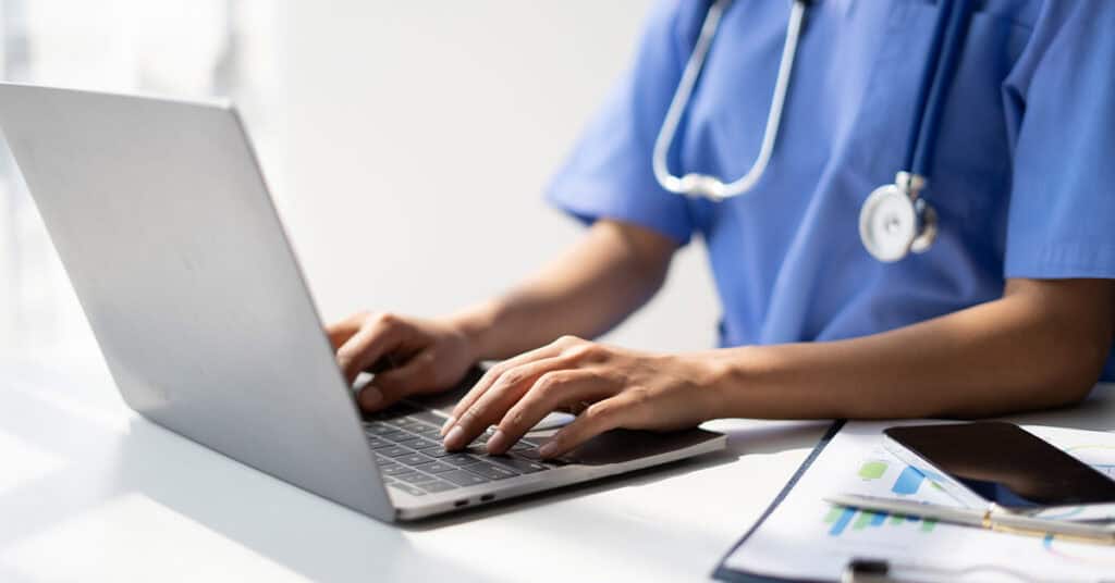 A person in blue medical scrubs types on a laptop at a desk with a stethoscope, clipboard with charts, and a smartphone nearby.