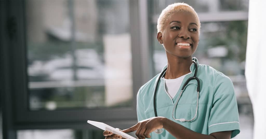 A nurse in scrubs with a stethoscope around her neck is smiling and holding a tablet, standing indoors near a window.