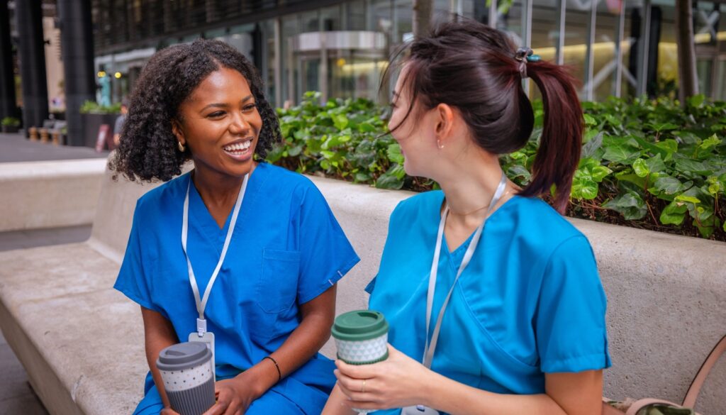 Two women in blue scrubs sit on a bench outdoors, holding reusable coffee cups and smiling at each other.