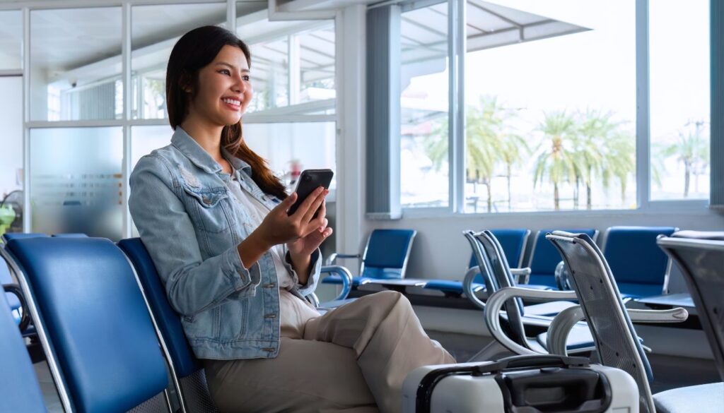 A woman sits in an airport waiting area, holding a smartphone and smiling, with a suitcase beside her and palm trees visible outside the window.