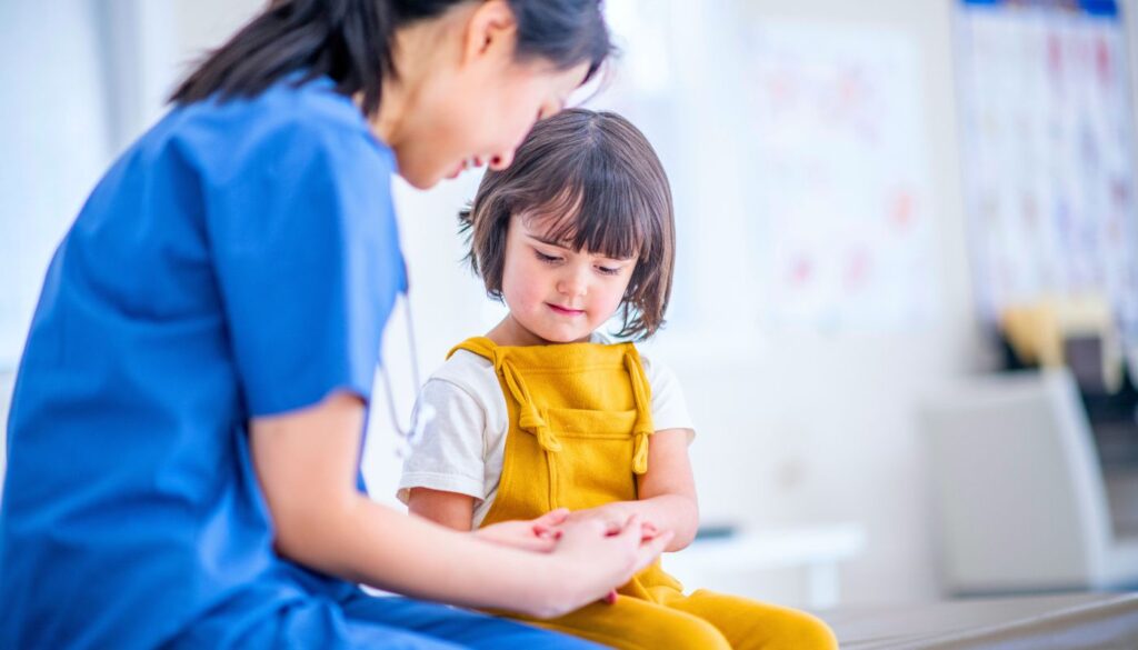 A nurse in blue scrubs holds hands with a young girl in yellow overalls, sitting together in a brightly lit medical setting.