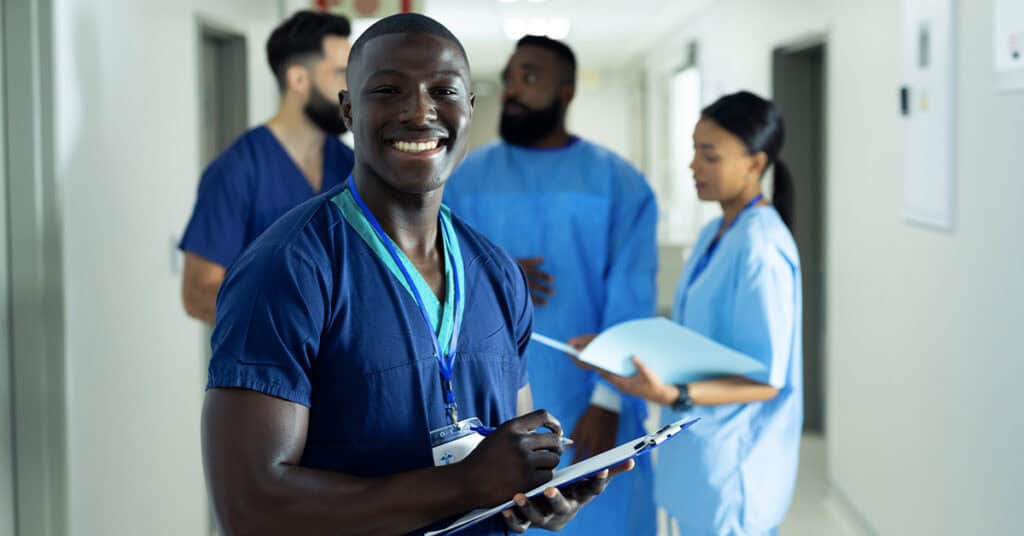 A healthcare worker in blue scrubs smiles while holding a clipboard in a hallway with three colleagues in the background.