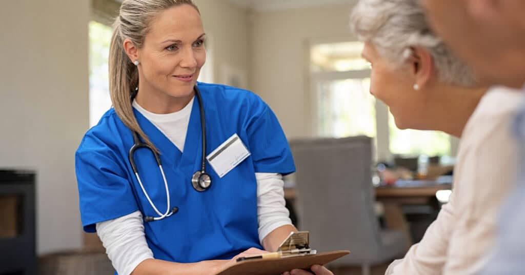 A nurse in blue scrubs with a stethoscope holds a clipboard and talks to an older adult in a well-lit room.