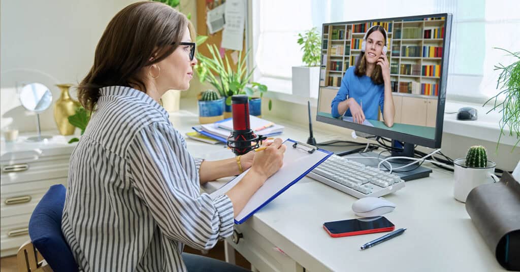 A woman sits at a desk taking notes while participating in a video call with another woman who appears on her computer screen.