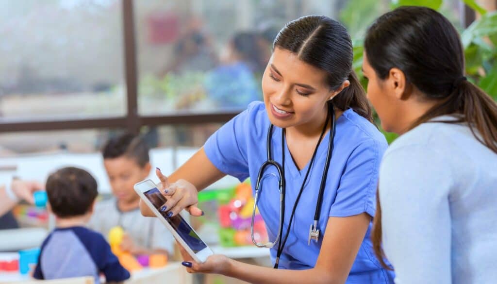 A healthcare professional in blue scrubs shows information on a tablet to another adult while children play in the background.