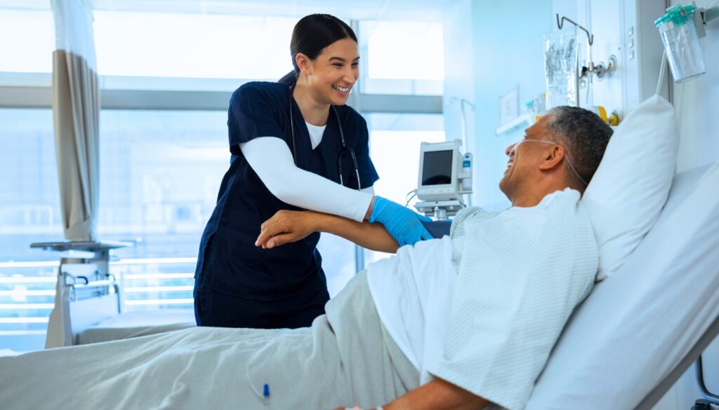 A nurse in navy scrubs smiles while checking a patient's arm as he lies in a hospital bed in a bright hospital room.
