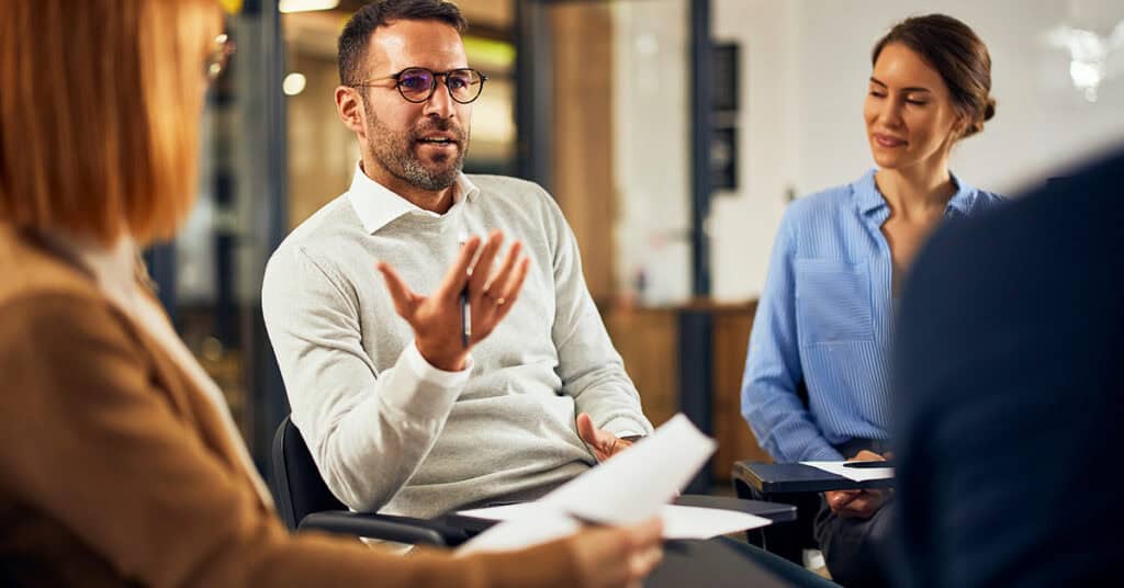 A man speaks while gesturing with his hand during a meeting, seated in a group with two women and another person, all holding papers.