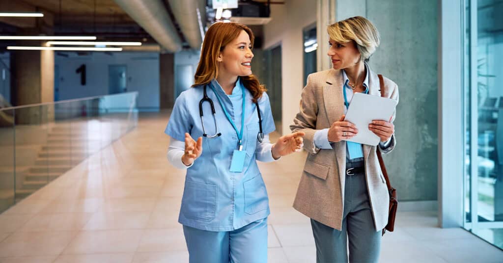 A nurse in scrubs talks with a professional woman holding a tablet as they walk together through a modern hallway.