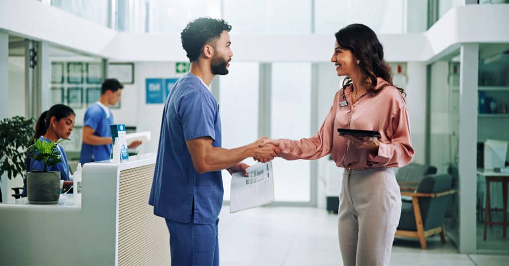 A man in medical scrubs and a woman with a tablet shake hands in a modern medical office reception area. Two other people work in the background.