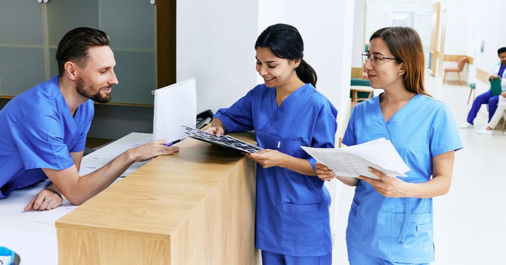 Three medical professionals in blue scrubs review documents together at a reception desk in a hospital or clinic setting.