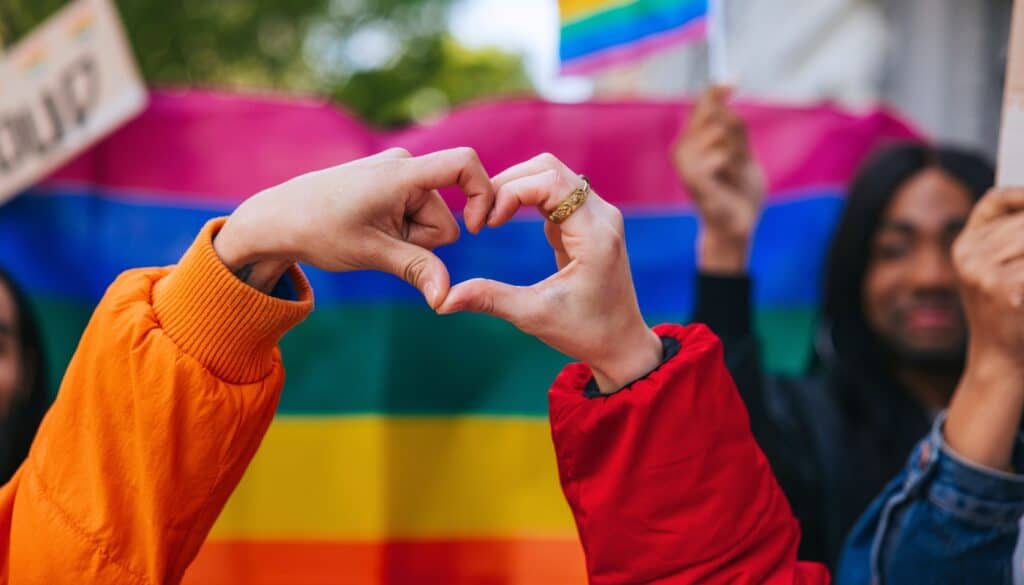 Two people form a heart shape with their hands in front of a rainbow Pride flag, with others holding signs in the background.