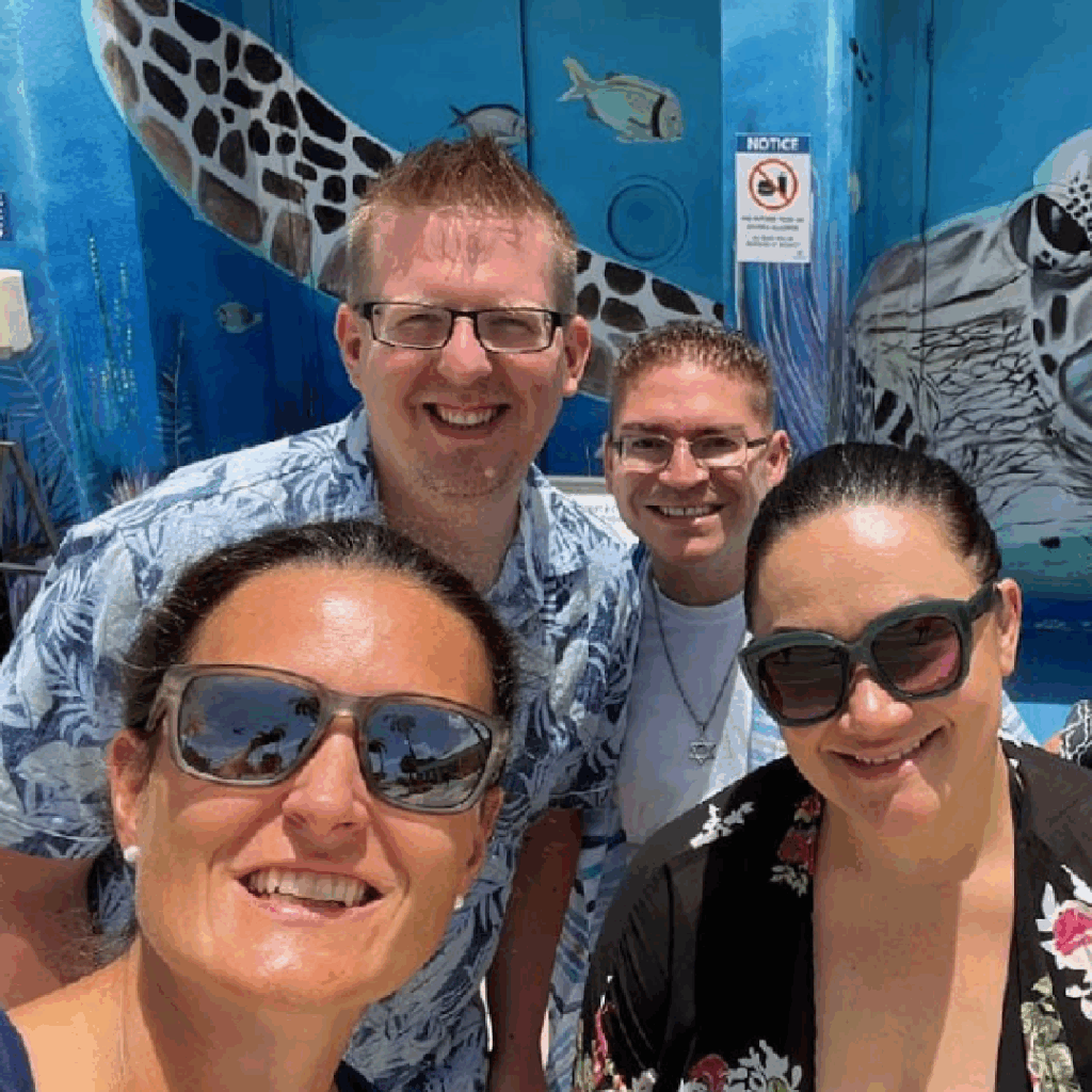 Four adults pose for a selfie outdoors in front of a colorful mural featuring a large sea turtle and fish.