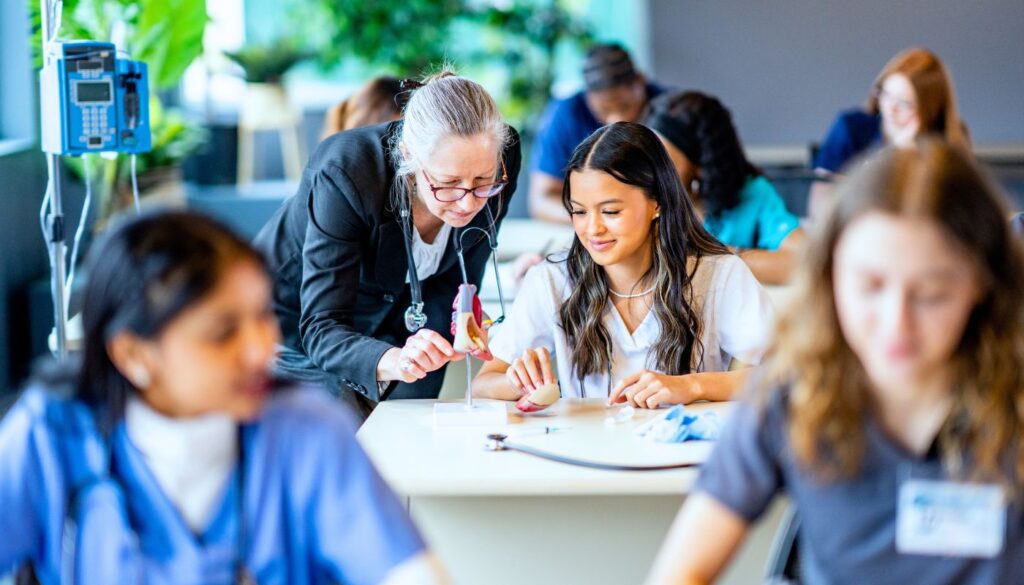 A teacher assists a student with a medical model in a classroom, while other students work at their desks in the background.