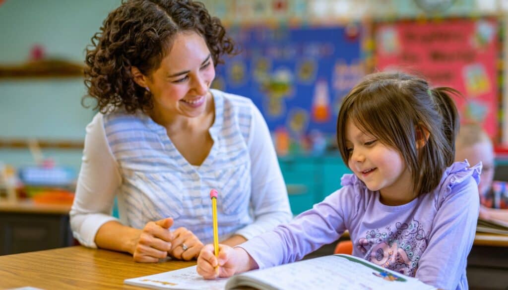 A young girl writes in a notebook while a woman, possibly a teacher, sits beside her and smiles in a colorful classroom setting.