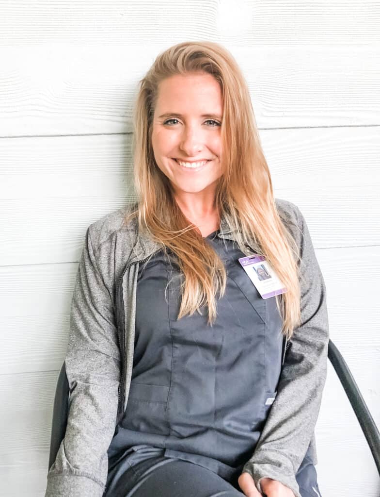 A woman with long blonde hair, wearing dark scrubs and a gray jacket, sits and smiles. She has an ID badge clipped to her jacket. The background is a light-colored wooden wall.