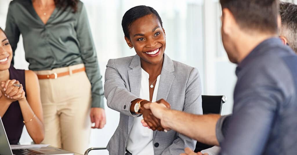 A woman in a business suit smiles and shakes hands with a man at a meeting table, while colleagues observe in a modern office setting.
