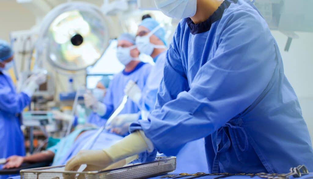 Medical staff in surgical gowns work in an operating room, with one person preparing surgical instruments and others attending to a patient in the background.