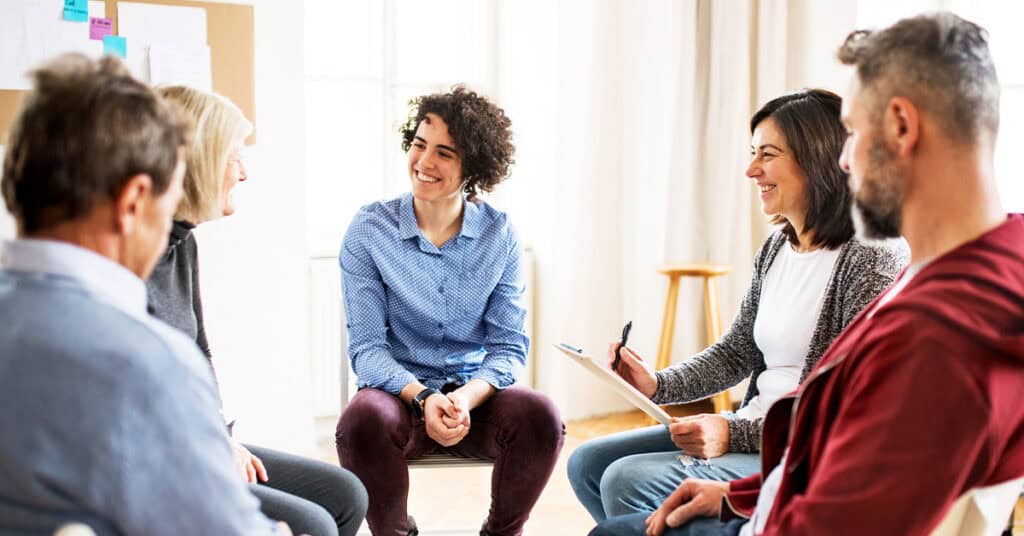 A group of five adults sit in a circle indoors, talking and smiling, as one person holding a clipboard and pen leads the discussion—a warm example of teamwork in behavioral health staffing.