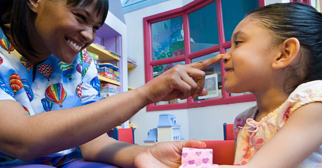An adult gently touches a smiling girl's nose in a colorful room, possibly a classroom or playroom, with toys and books visible on shelves in the background.