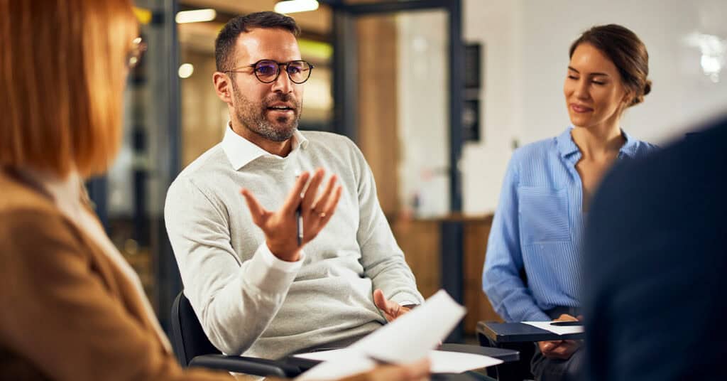 A man in glasses speaks while seated in a group discussion with three others in a modern office setting.