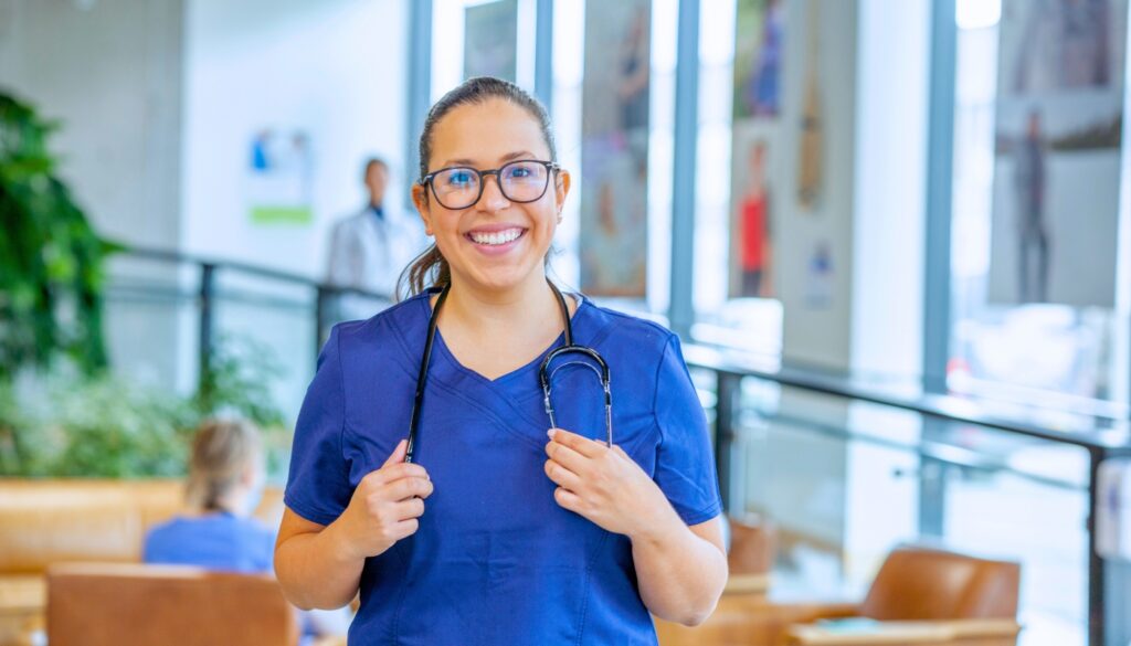 A woman in blue medical scrubs and glasses stands indoors, smiling and holding a stethoscope around her neck. The background shows a modern, well-lit space with people and plants.