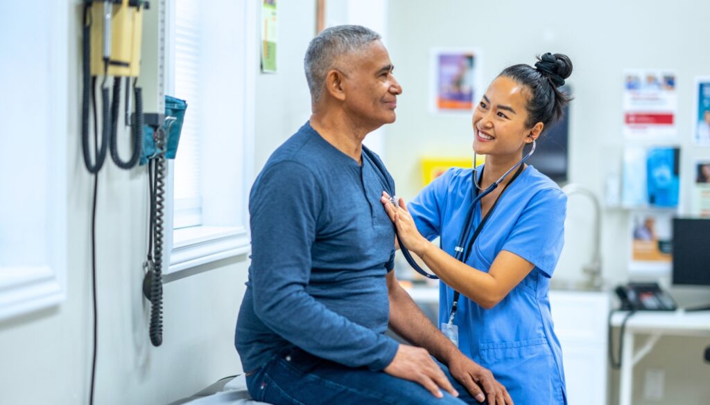 A nurse wearing blue scrubs uses a stethoscope to check the chest of a seated man during a medical examination in a clinic room.