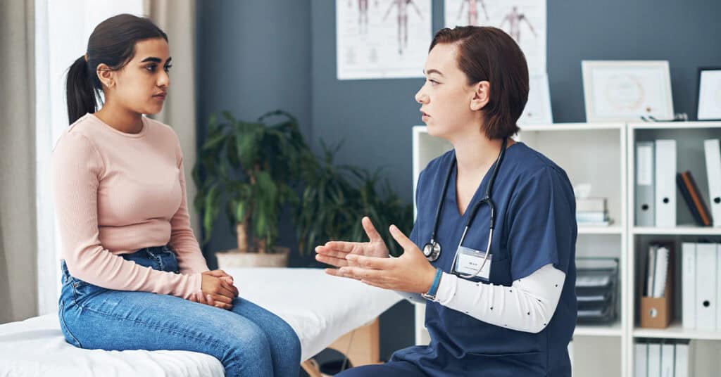 A doctor in scrubs speaks to a seated patient in a medical office. The patient listens attentively. Medical charts and office supplies are visible in the background.