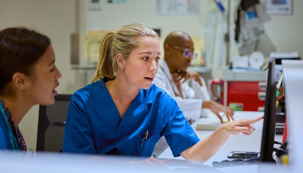 Two healthcare professionals in scrubs look at a computer monitor while another person works at a desk in the background.