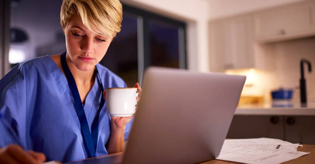 A person in scrubs sits at a table with a laptop and paperwork, holding a mug and appearing focused on their work.
