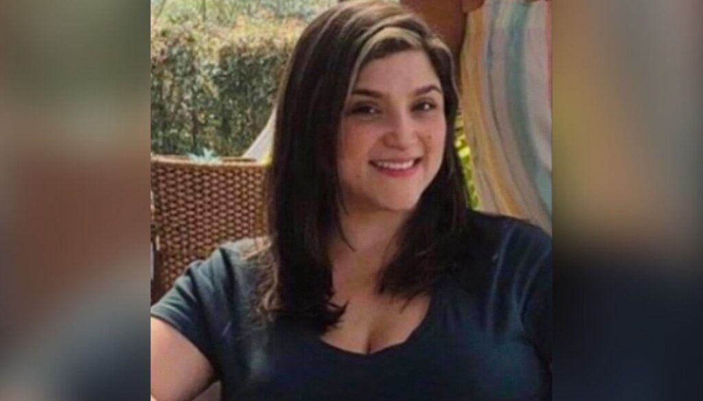 A woman with shoulder-length dark hair, wearing a dark top, sits and smiles at the camera outdoors.