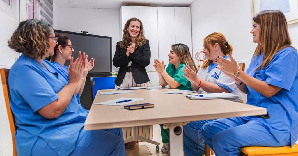 A group of healthcare professionals in scrubs clap while a woman in business attire stands and smiles at the front of a meeting table.