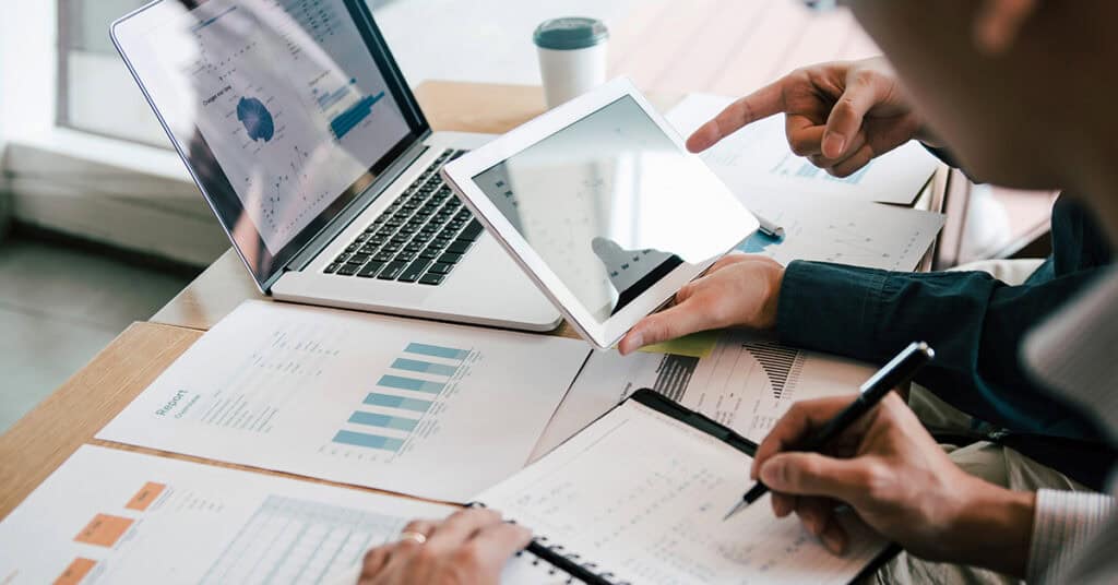 Two people review charts and graphs on papers, a laptop, and a tablet at a desk, discussing data analysis or business reports.