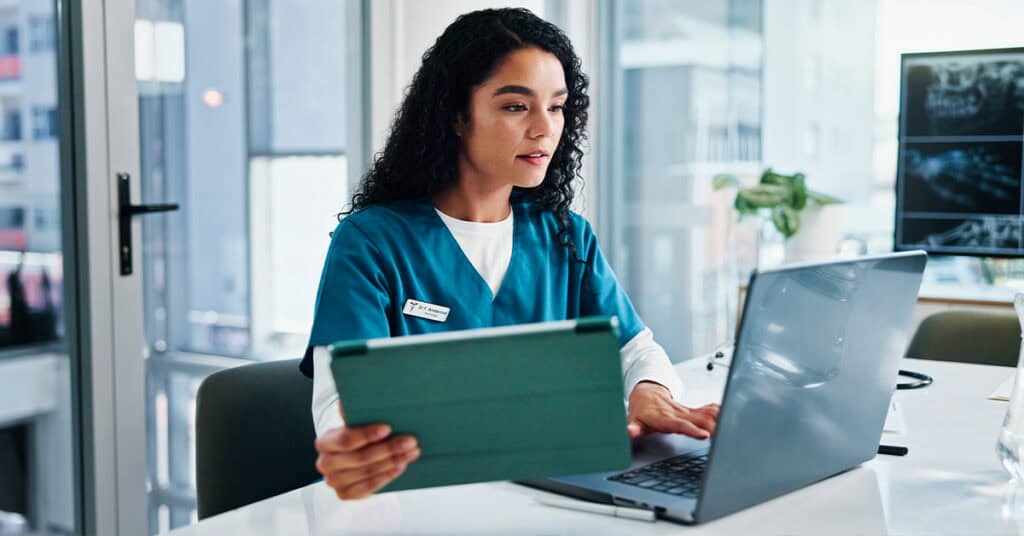 A woman in medical scrubs sits at a desk, holding a clipboard and working on a laptop in a modern office.