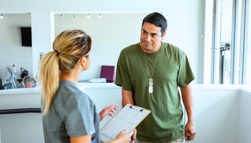 A woman holding a clipboard talks to a man wearing a green t-shirt and dog tags in a bright indoor setting.