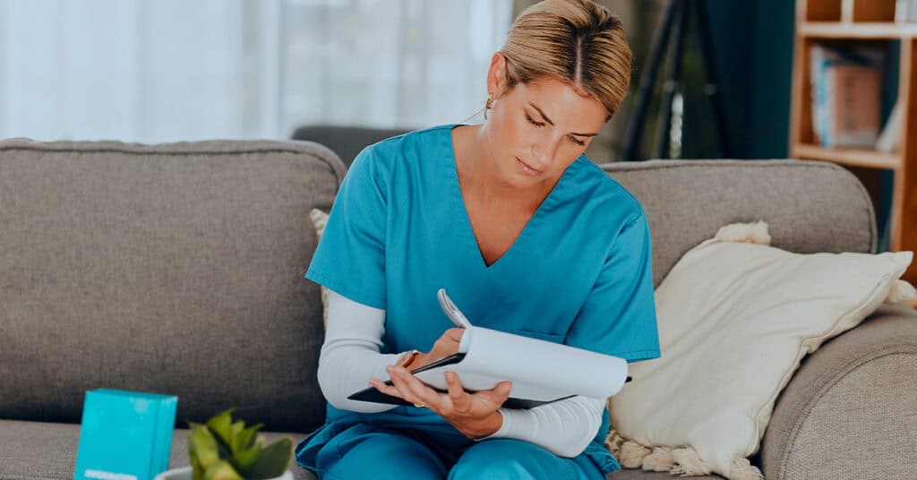 A person in blue medical scrubs sits on a couch, writing in a notebook with a pen.