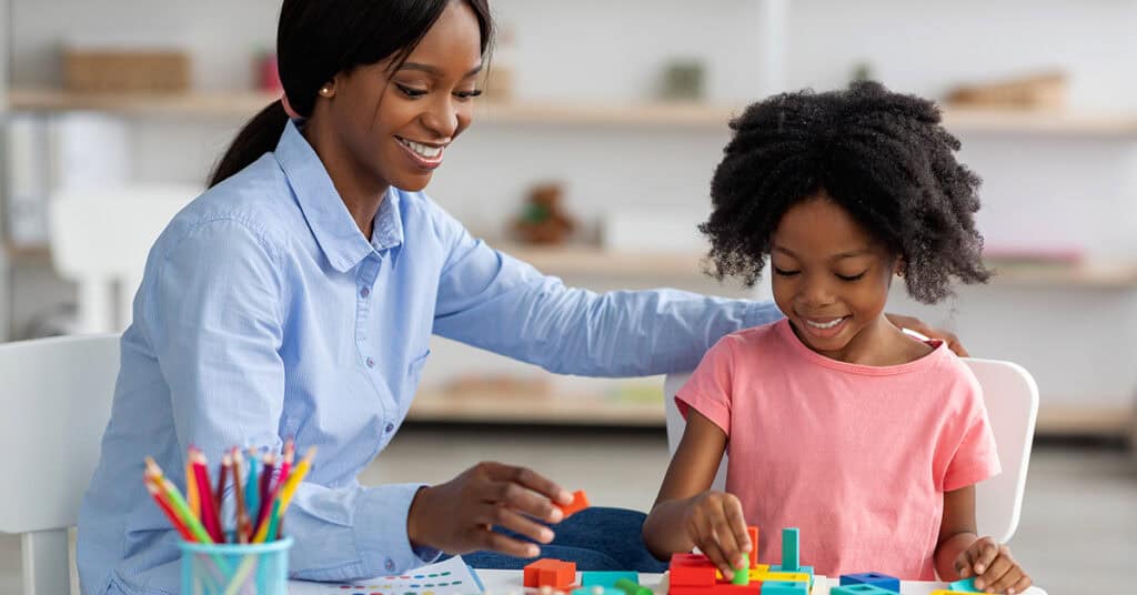 An adult and a child sit at a table together, smiling and playing with colorful building blocks. A cup of colored pencils is visible in the foreground.