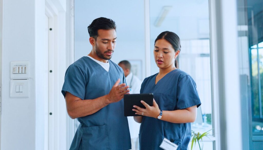 Two healthcare professionals in scrubs discuss information displayed on a tablet in a medical facility hallway.