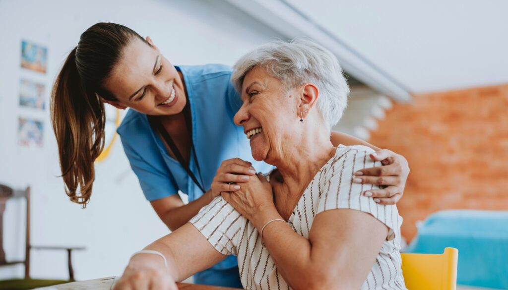 A healthcare worker in blue scrubs smiles and gently supports an older woman seated at a table, who is also smiling.