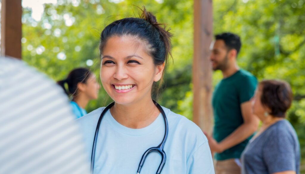 A smiling healthcare worker with a stethoscope stands outdoors, with other people conversing in the blurred background on a sunny day.