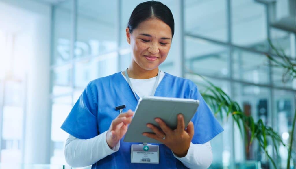 A travel nurse in blue scrubs uses AI on a tablet device while standing in a bright, modern medical facility.