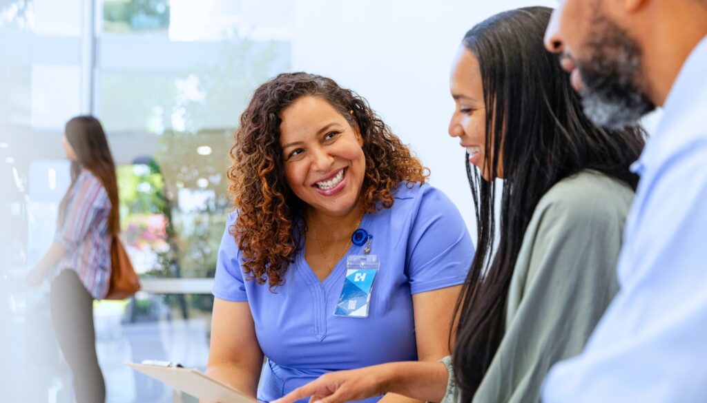A nurse in scrubs smiles and holds a clipboard while talking with two African American adults seated beside her in a brightly lit room.