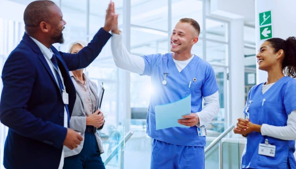 Two healthcare workers in scrubs high-five each other while smiling, with a third colleague and another person in business attire observing in a hospital hallway.