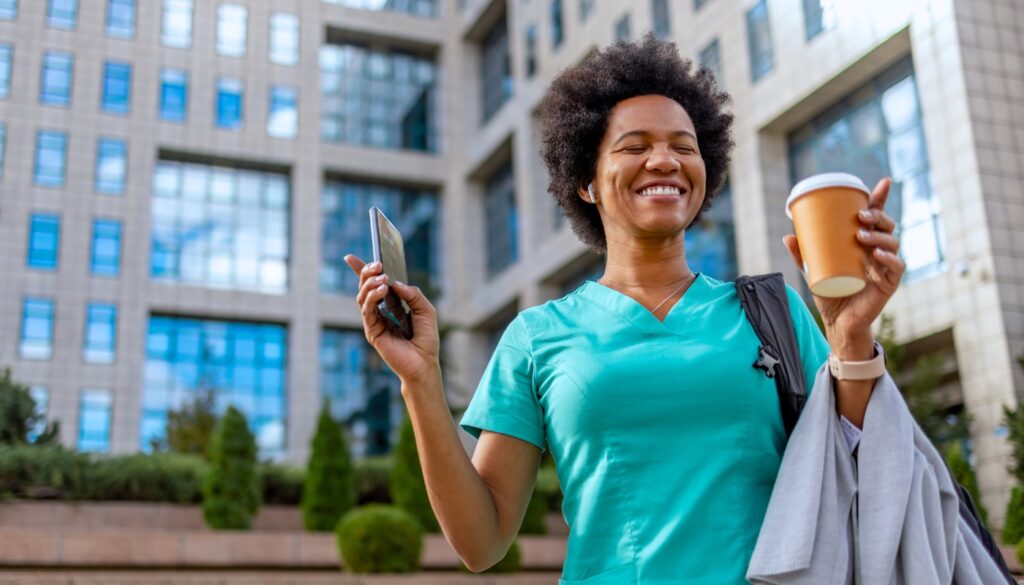 Smiling travel nurse in scrubs stands outside a modern building, holding a smartphone, coffee cup, and jacket.