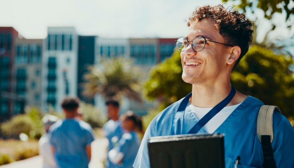 A young travel nurse in blue medical scrubs smiles outdoors, holding a folder and wearing a lanyard, with other people in scrubs and buildings in the background.