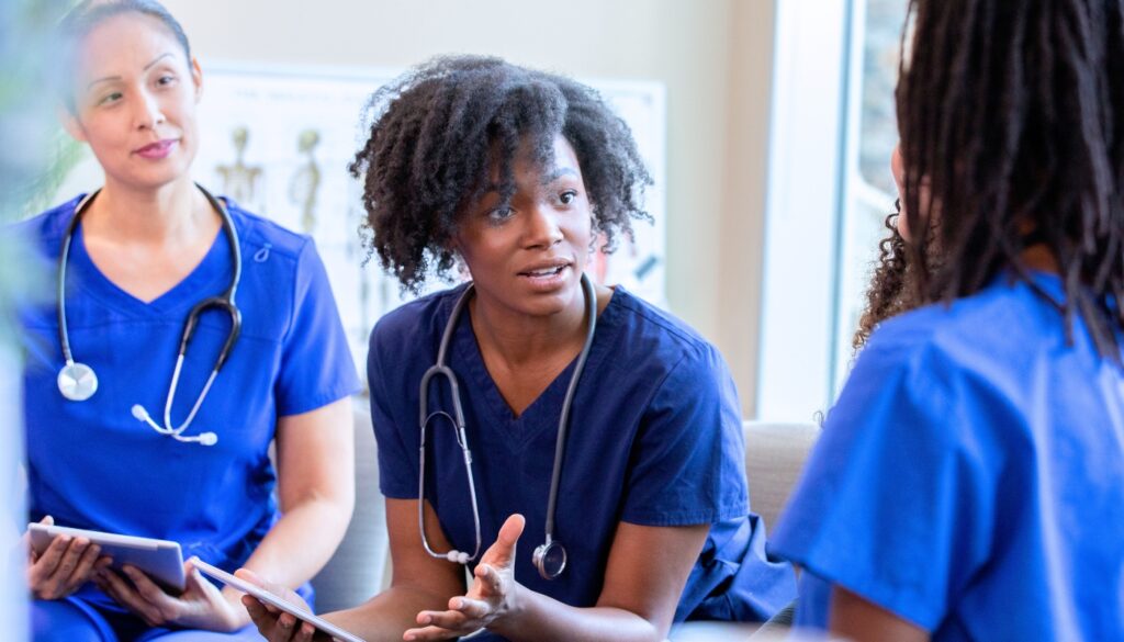 Three travel nurses in blue scrubs, with stethoscopes, sit together having a discussion about mental health indoors.